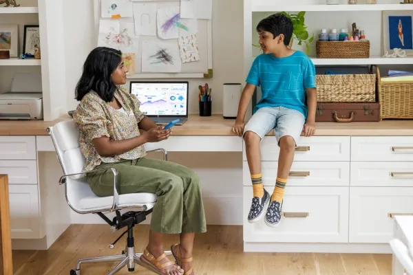 A mother sits in a white office chair looking at her son, who is sitting on a white and wood desk beside a laptop displaying charts and graphs.